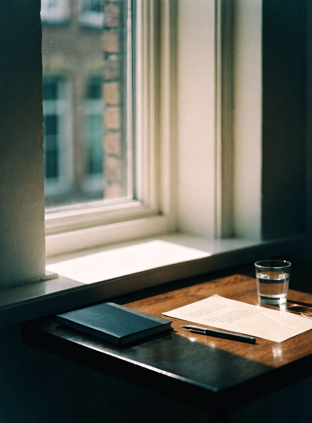 Un escritorio junto a la ventana, con un cuaderno cerrado y una lapicera.